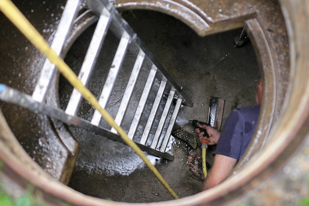 Cleaning technician utilizes a hydro jet tool to clean scum from the surface of a sewage line by climbing down with a ladder. 